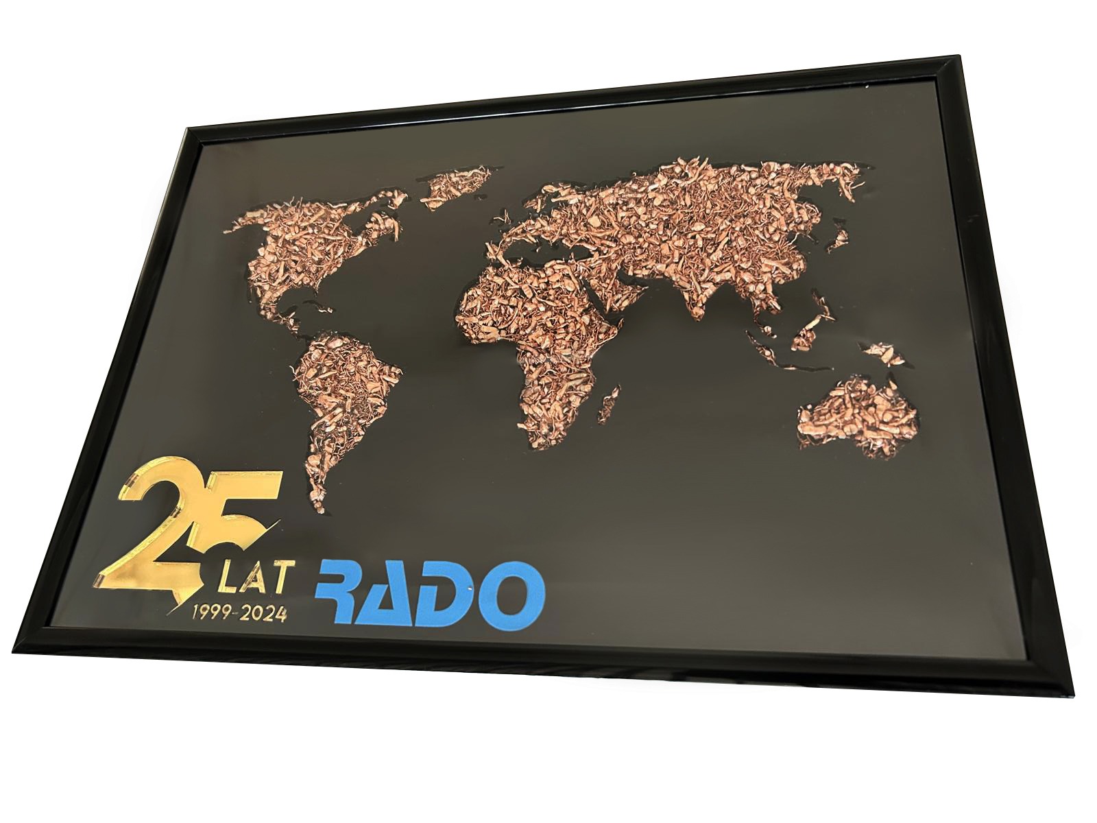 Oprawiona w ramę mapa świata wykonana z drobnych elementów w odcieniach brązu na czarnym tle, z napisem '25 LAT RADO 1999-2024' w kolorach złotym i niebieskim.
