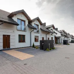 Nowoczesne domy szeregowe w Gnojniku z drewnianymi drzwiami wejściowymi i oknami, widok z brukowanej drogi dojazdowej. Eleganckie wykończenie.