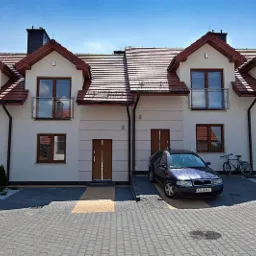 Elewacja szeregowca z brązowymi oknami i drzwiami, z balkonami z balustradami ze szkła. Na podjeździe zaparkowany samochód, obok stoi rower.