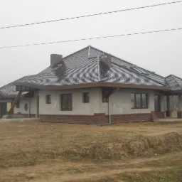 Dom jednorodzinny z szarą dachówką karpiówką, widoczne okna dachowe i ceglany komin, w tle mglista pogoda.