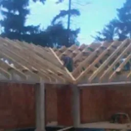 Konstrukcja drewniana więźby dachowej w trakcie montażu, widoczny pracownik na szczycie, surowy stan, ceglane ściany budynku w tle.