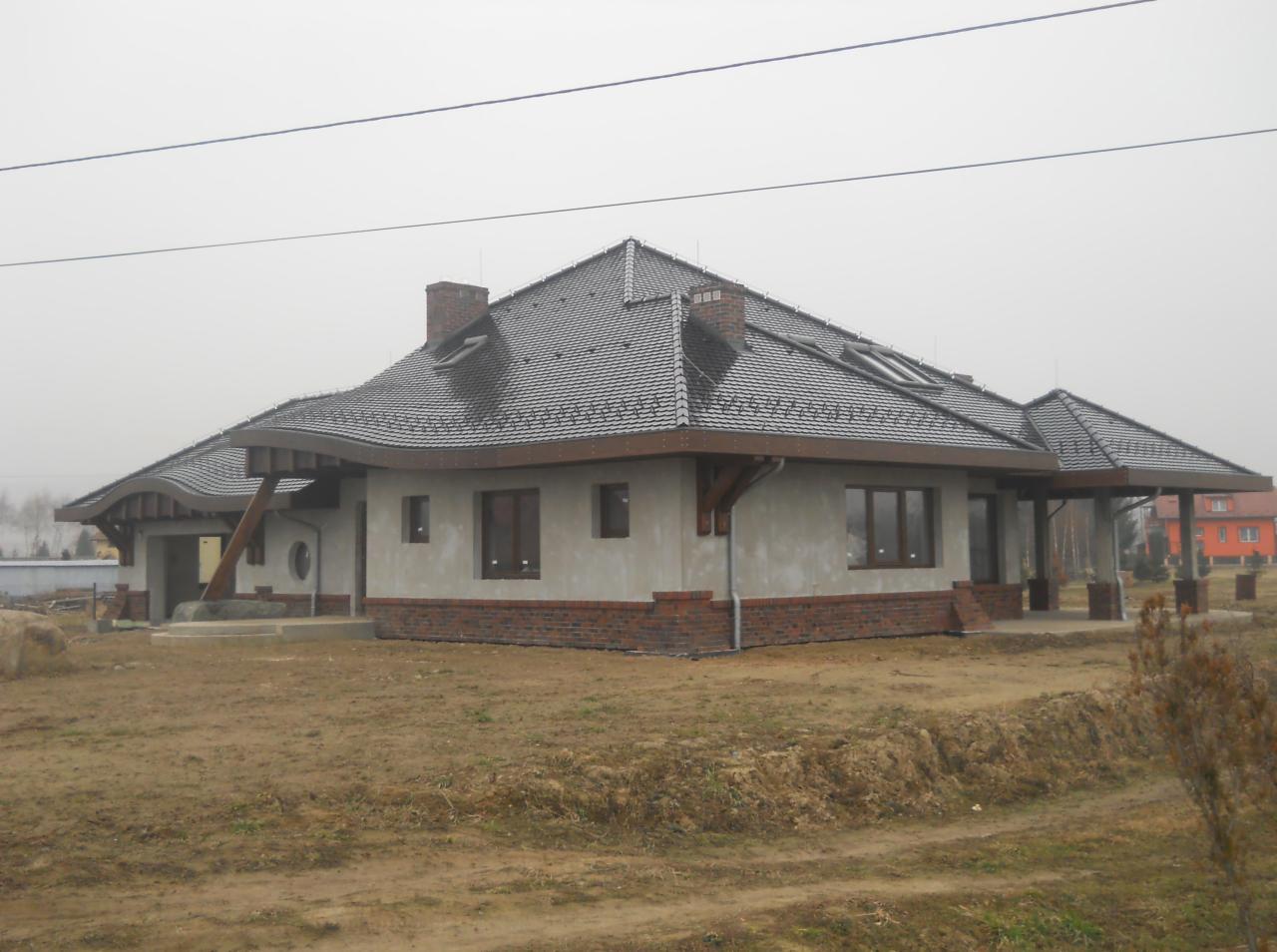 Dom jednorodzinny z szarą dachówką karpiówką, widoczne okna dachowe i ceglany komin, w tle mglista pogoda.