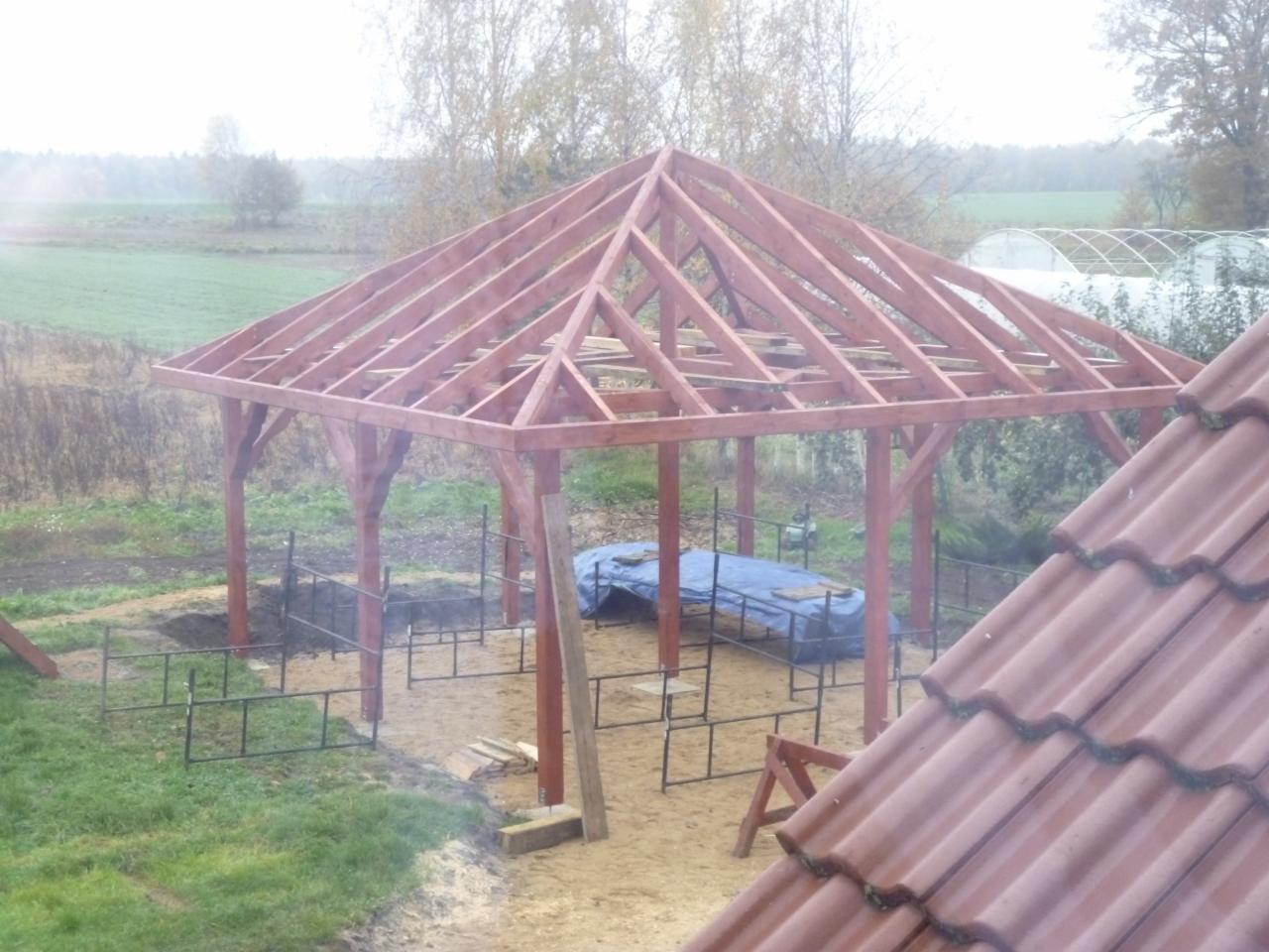 Wznoszona konstrukcja drewniana altany z widoczną więźbą dachową, częściowo zadaszona, na tle zarośniętego terenu i fragmentu dachu z dachówki ceramicznej.