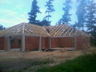 Budowa domu w stanie surowym otwartym: widoczna murowana konstrukcja ścian i drewniana więźba dachowa bez pokrycia, z elementami betonowych słupów.