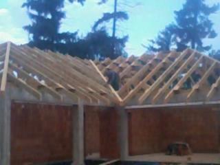 Konstrukcja drewniana więźby dachowej w trakcie montażu, widoczny pracownik na szczycie, surowy stan, ceglane ściany budynku w tle.