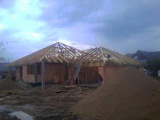 Wznoszenie drewnianej konstrukcji dachu domu jednorodzinnego, widoczne krokwie, murłaty i słupy, w tle zachmurzone niebo.