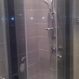 Wykończona kabina prysznicowa z szarymi płytkami i chromowaną armaturą. Widoczne oświetlenie punktowe w płytkach i fragment toalety. Nowoczesny design z dbałością o detale.