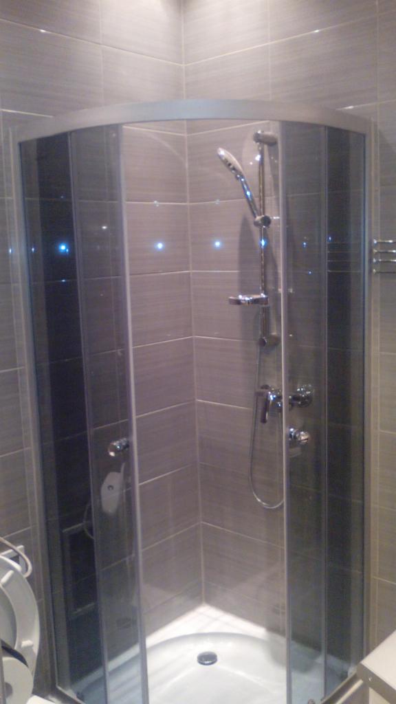 Wykończona kabina prysznicowa z szarymi płytkami i chromowaną armaturą. Widoczne oświetlenie punktowe w płytkach i fragment toalety. Nowoczesny design z dbałością o detale.