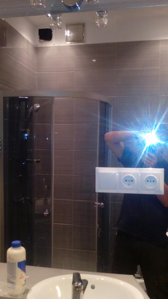 Wykończona łazienka w Krakowie z kabiną prysznicową i nową armaturą. Widoczne płytki na ścianach i biała umywalka z baterią. Ujęcie w lustrze z odblaskiem.