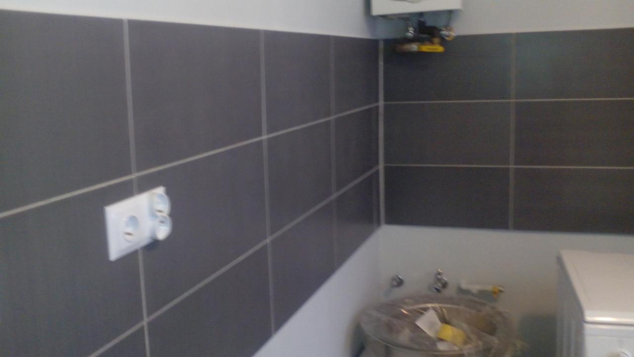 Szare płytki z fugami na ścianie w łazience, widoczne gniazdko elektryczne i fragment umywalki. Ujęcie z bliska, prezentujące wykończenie wnętrza.