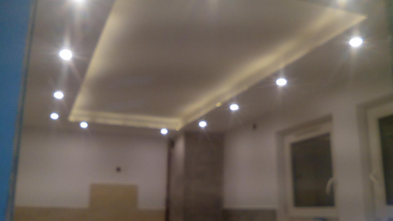 Wykończony sufit podwieszany z oświetleniem punktowym w jasnym wnętrzu. Widoczne okno i fragment ściany. Delikatne światło podkreśla nowoczesny design.