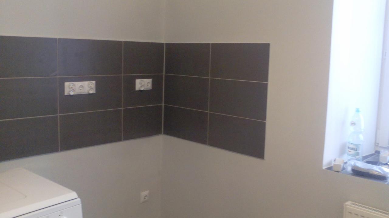 Ciemne, kwadratowe płytki na ścianie w rogu łazienki, z widocznymi gniazdkami elektrycznymi, nad pralką. Ściany pomalowane na szaro, okno z butelką w tle.