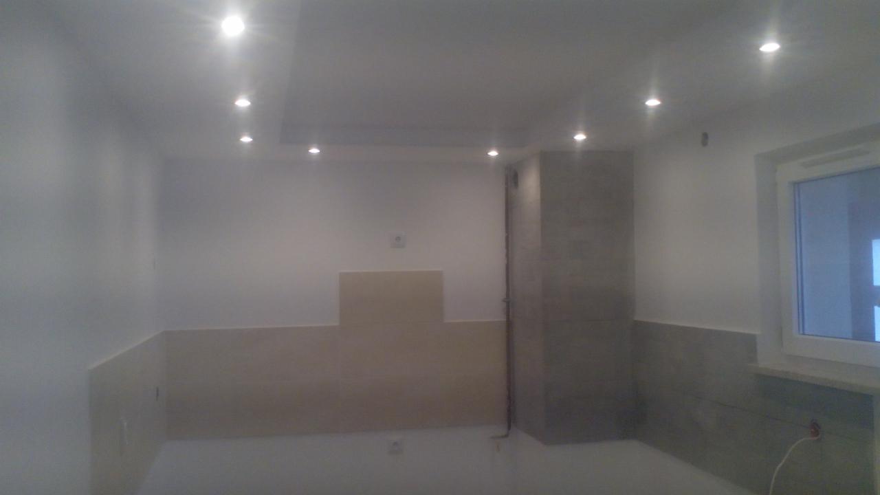 Wnętrze w trakcie remontu: białe ściany, częściowo położone płytki w odcieniach beżu i szarości. Sufit z oświetleniem punktowym. Okno po prawej stronie.