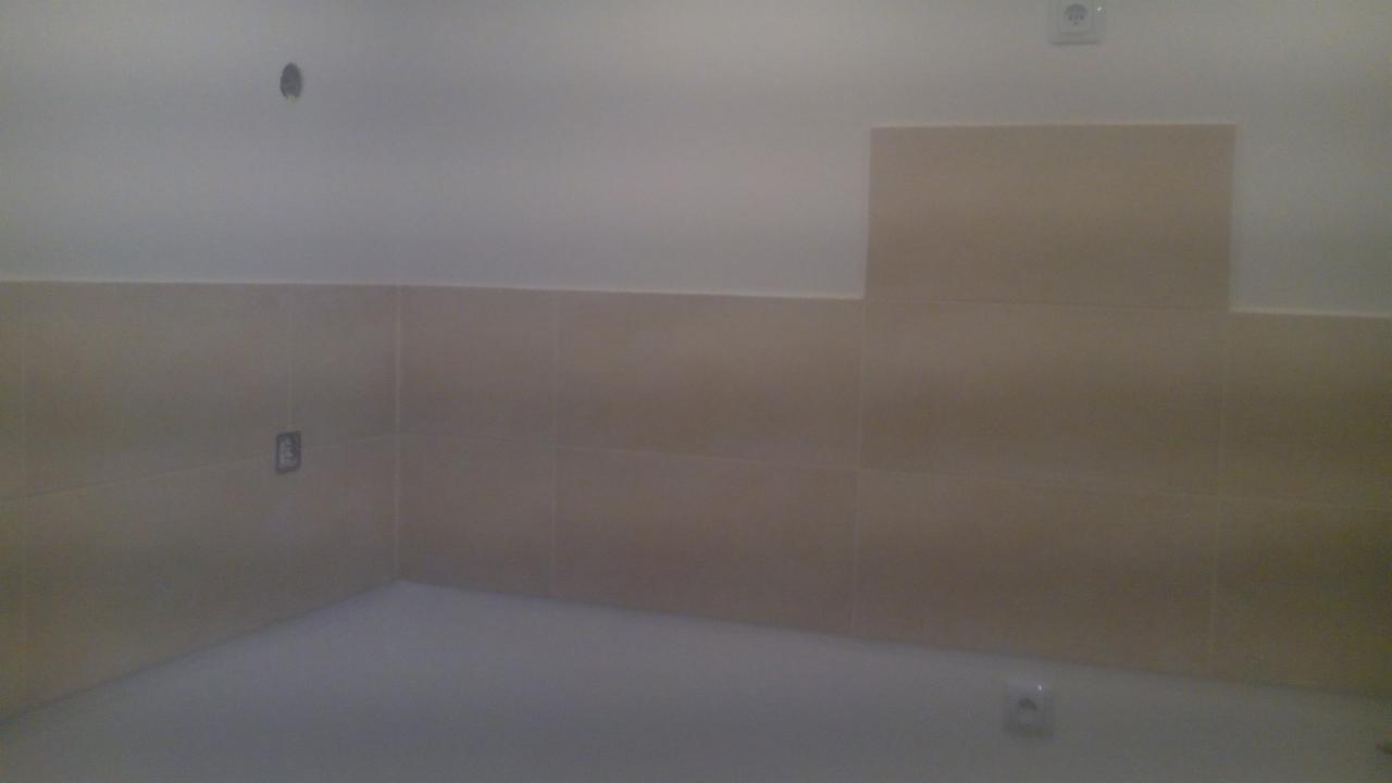 Beżowe płytki ścienne w łazience, częściowo ułożone, z widocznymi gniazdkami elektrycznymi i otworem na rurę, na tle białej ściany.