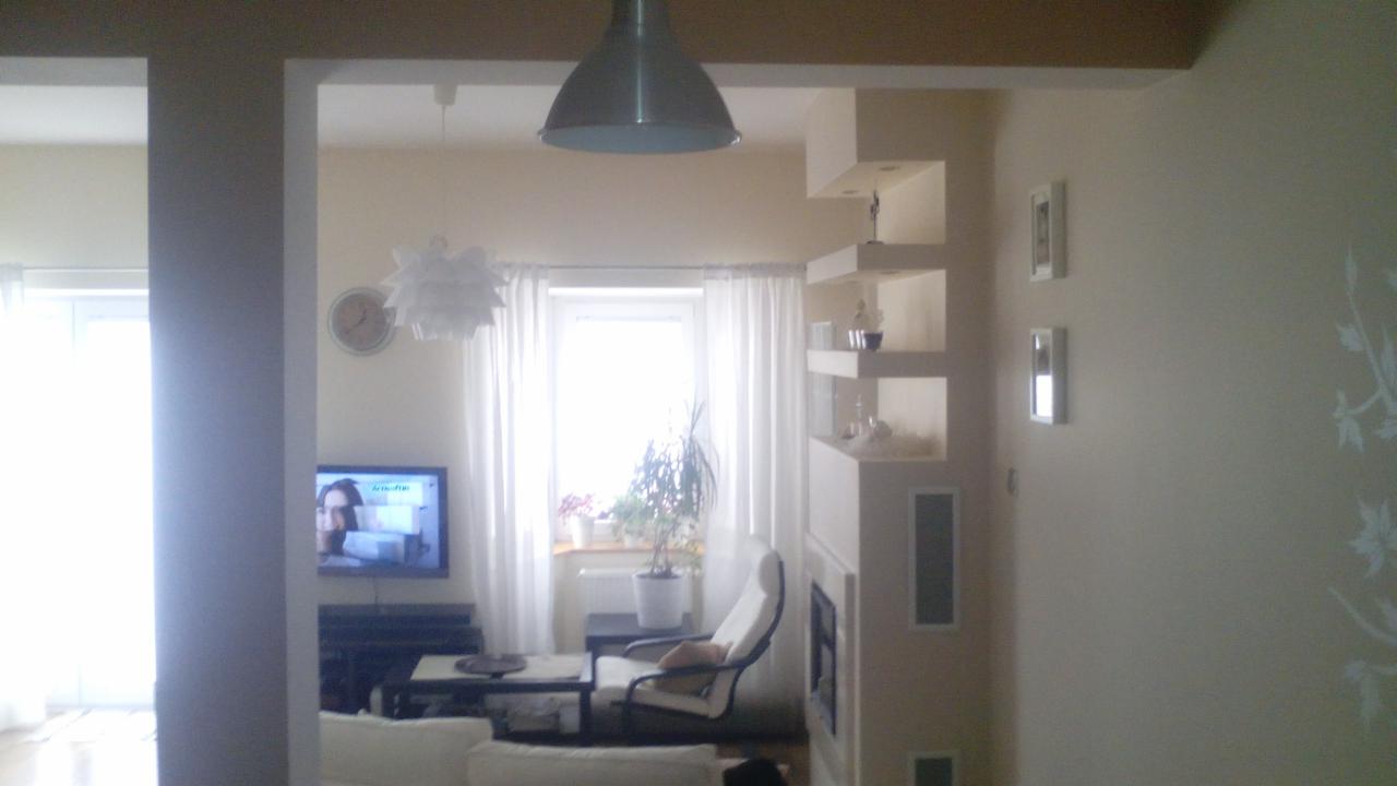 Jasne wnętrze po remoncie: kremowe ściany z delikatnym wzorem, designerski regał i lampa wisząca. Widok na salon z telewizorem i oknem.