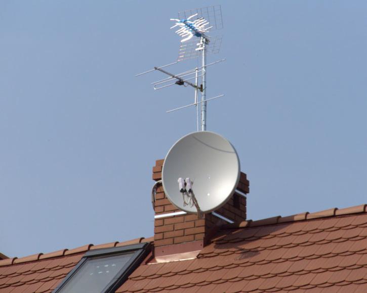 Antena satelitarna i antena naziemna zamontowane na kominie na dachu z dachówką ceramiczną i oknem dachowym, na tle błękitnego nieba.