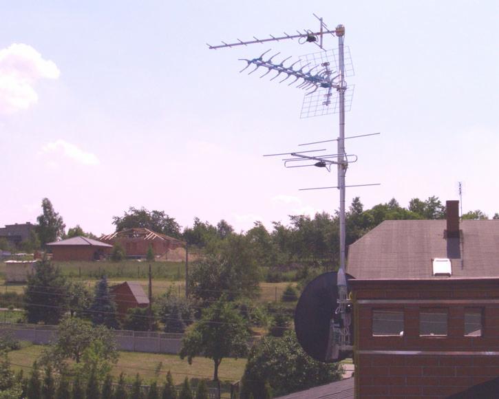 Złożona antena telewizyjna zamontowana na dachu budynku z czerwonej cegły, na tle zieleni i zabudowań. Widoczna antena satelitarna. Ujęcie z dołu.