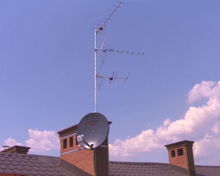 Zestaw antenowy na dachu z dachówki i ceglanym kominem, na tle błękitnego nieba. Antena satelitarna i kilka anten kierunkowych.