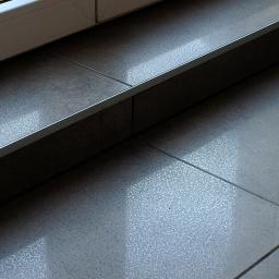 BUD-SYSTEM Marek Bojczuk - Szare płytki podłogowe przy progu okna, wykończone srebrną listwą, z widocznymi smugami wody.