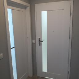 BUD-SYSTEM Marek Bojczuk - Dwie białe, nowoczesne drzwi wewnętrzne z pionowymi, matowymi przeszkleniami, zamontowane w szarym pomieszczeniu na szarej podłodze.