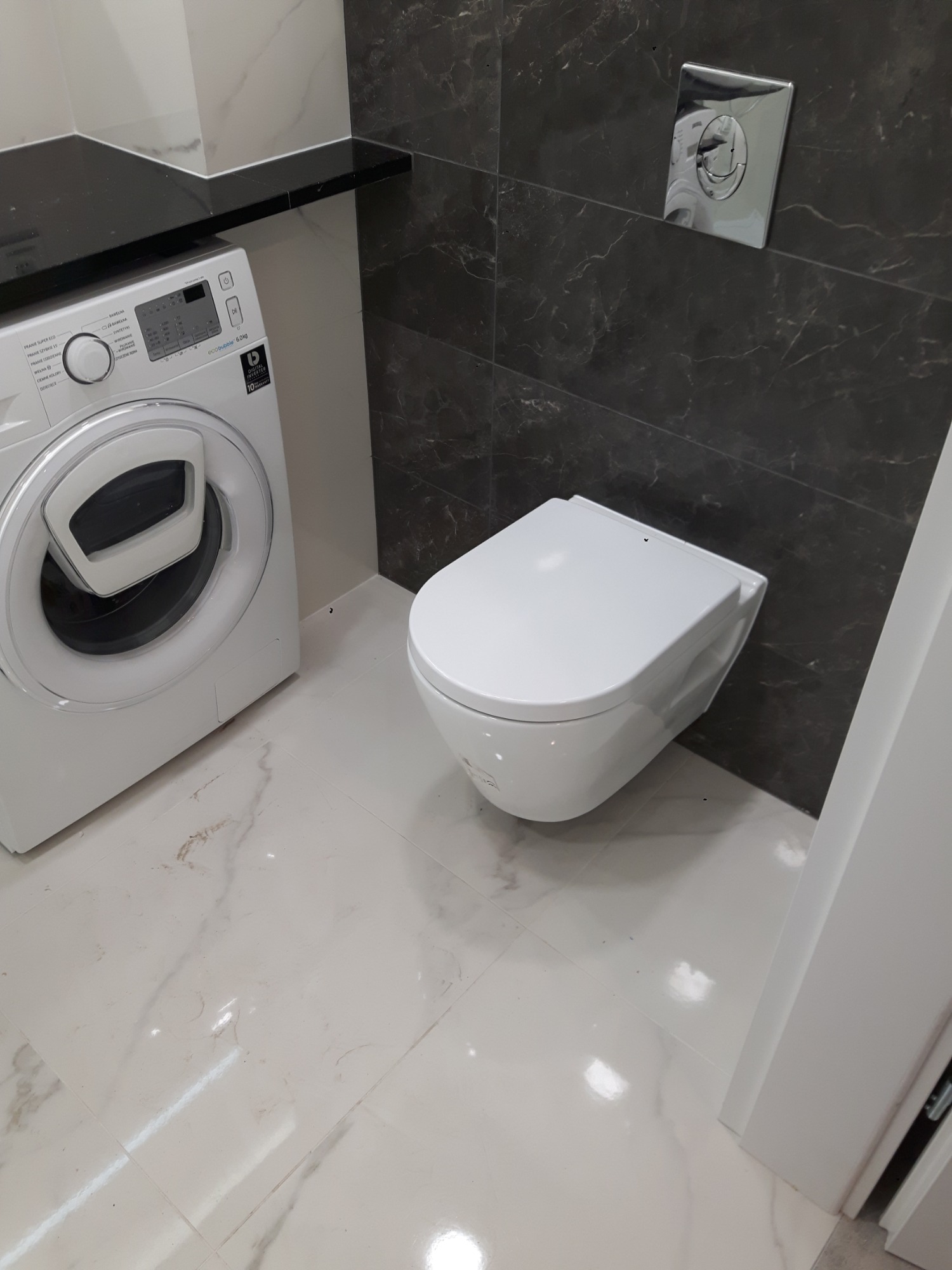 Nowoczesna łazienka z podwieszaną toaletą, pralką automatyczną i ciemnymi płytkami imitującymi marmur na ścianie, uzupełniona jasnymi płytkami podłogowymi z delikatnym wzorem.
