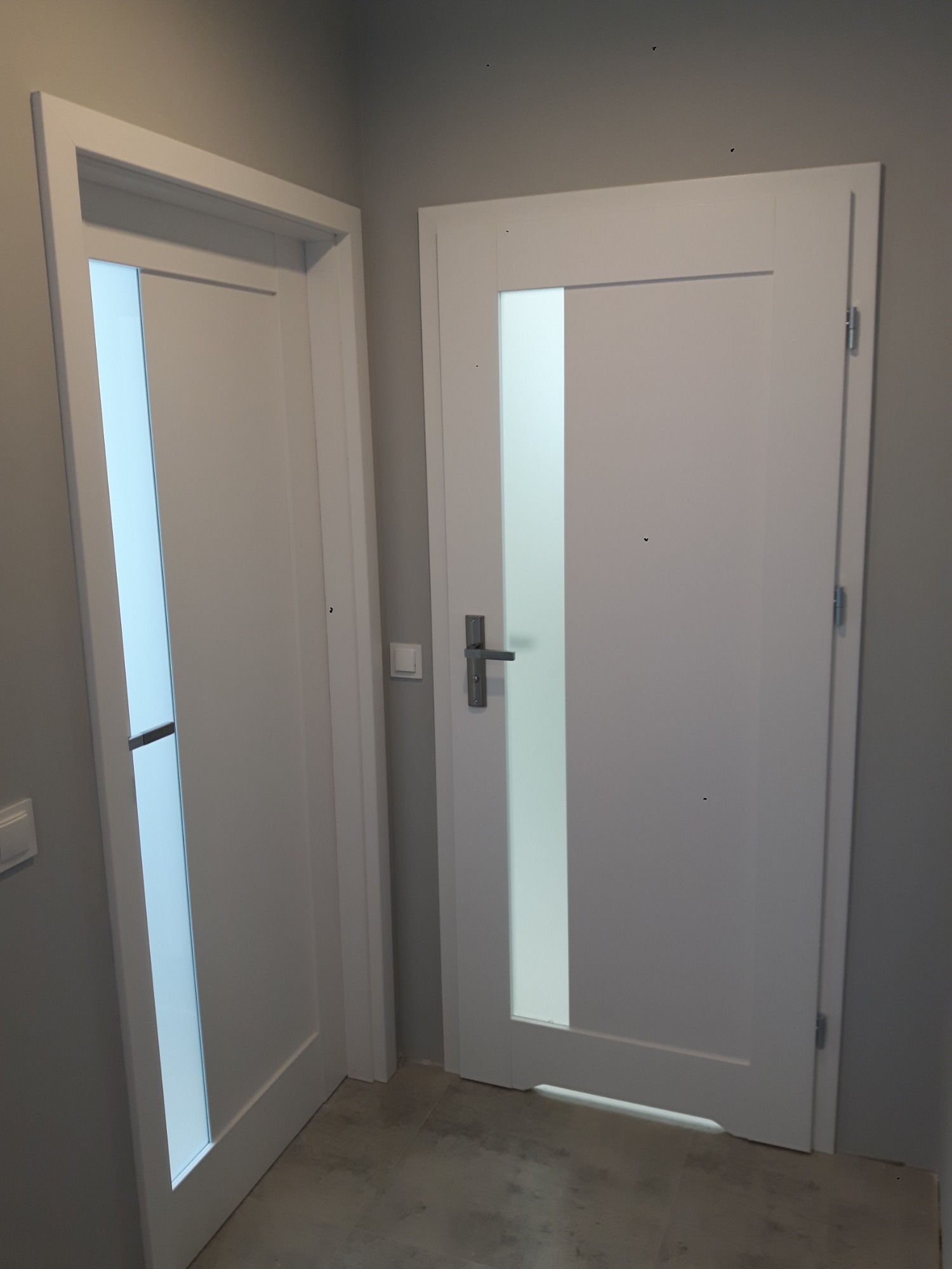 Dwie białe, nowoczesne drzwi wewnętrzne z pionowymi, matowymi przeszkleniami, zamontowane w szarym pomieszczeniu na szarej podłodze.