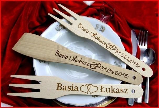 Drewniane sztućce (widelec i łopatka) z grawerem imion Basia i Łukasz oraz datą 31.06.2015, leżące na białym talerzu na tle czerwonego aksamitu.