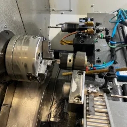 Precyzyjna obróbka metalu na tokarce CNC: zbliżenie na uchwyt z zamocowanym elementem i narzędzia skrawające w trakcie pracy, widoczne chłodziwo i wióry.