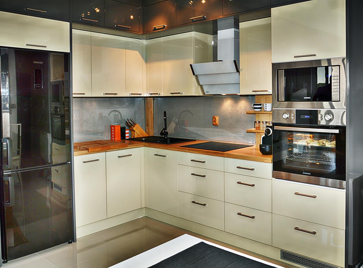 Nowoczesna kuchnia w kształcie litery L z drewnianymi blatami, beżowymi szafkami i czarnymi akcentami. Widoczny sprzęt AGD i oświetlenie podszafkowe.