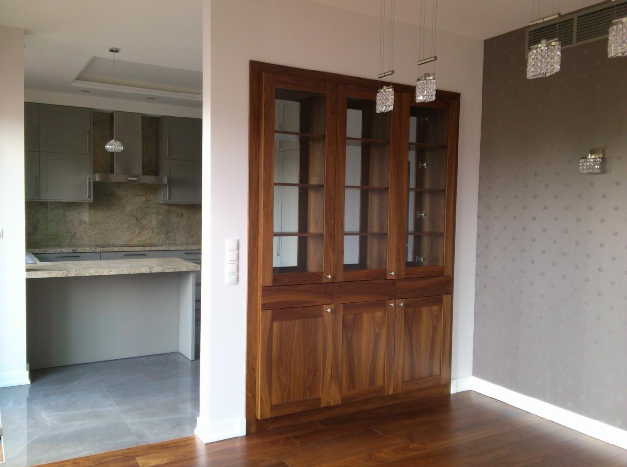 Drewniana szafa z przeszkleniami, wkomponowana we wnękę, widoczna z perspektywy otwartej kuchni w Raszynie. Ciepłe oświetlenie podkreśla naturalny rysunek drewna.