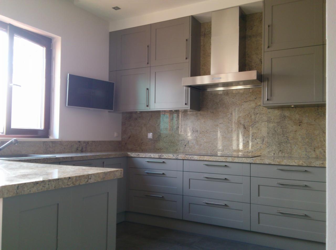 Szara kuchnia z blatem i ścianą z kamienia. Nad blatem szafki i okap. Na ścianie obok okna wisi telewizor. Nowoczesny design.