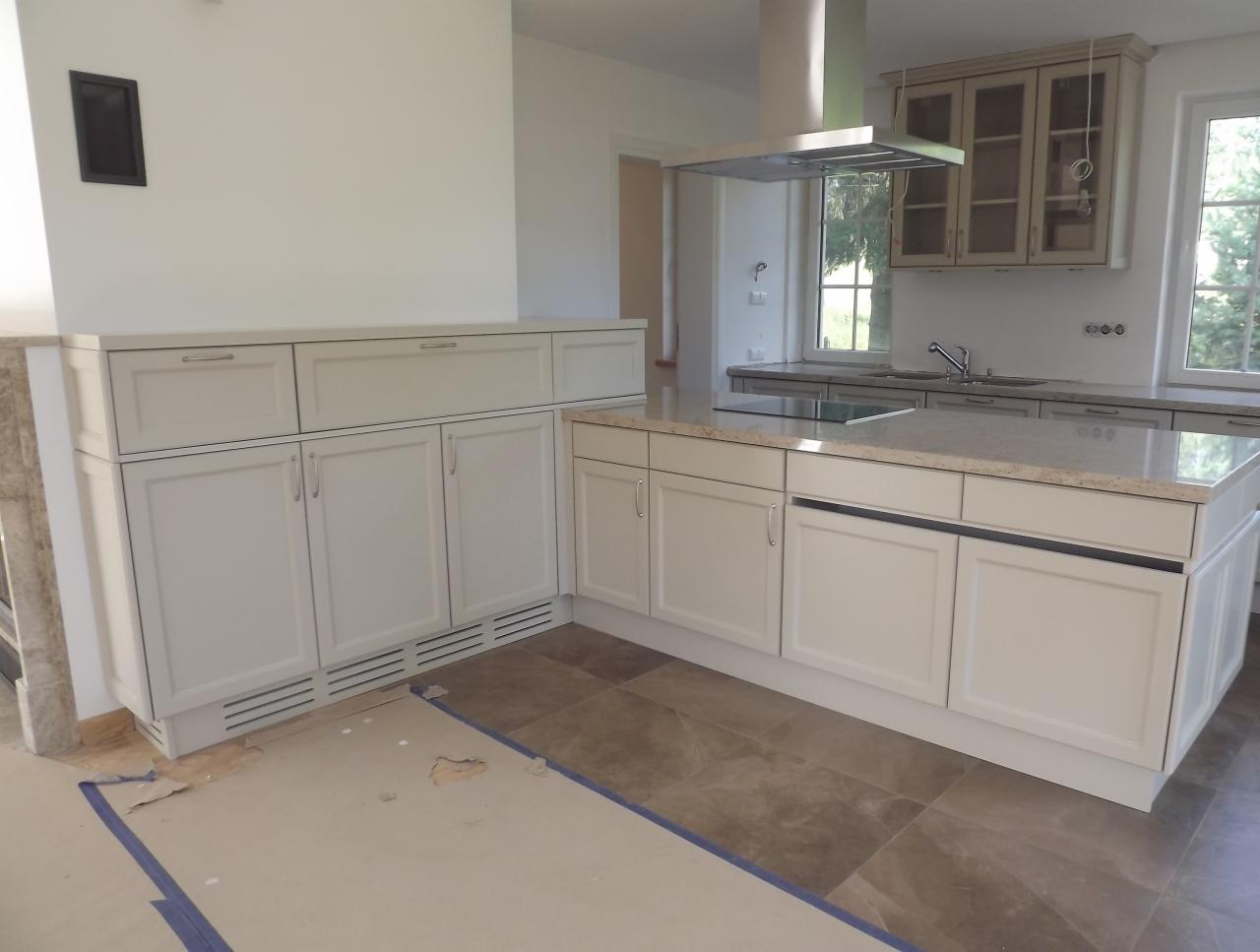 Jasna kuchnia z wyspą i zabudową pod oknem, w trakcie montażu. Widoczne zabezpieczenia podłogi i brakujące elementy wykończenia. Klasyczny styl.