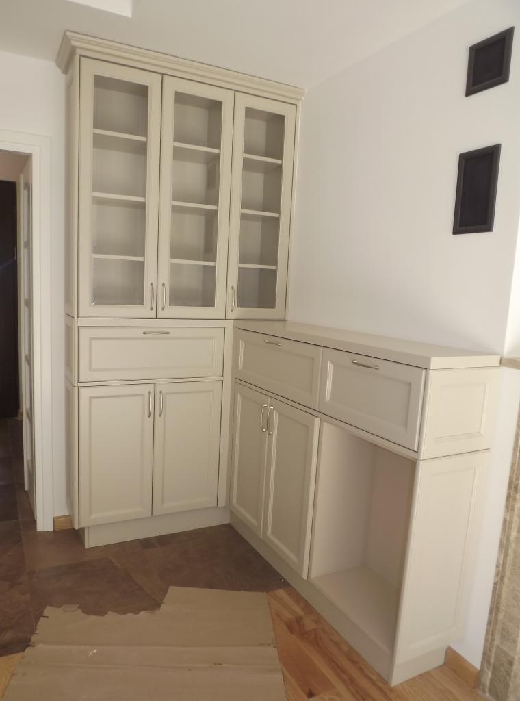 Jasna zabudowa kuchenna z przeszklonymi szafkami górnymi i dolnymi, widoczna w rogu pomieszczenia. Dwa czarne, prostokątne elementy dekoracyjne na ścianie w tle.