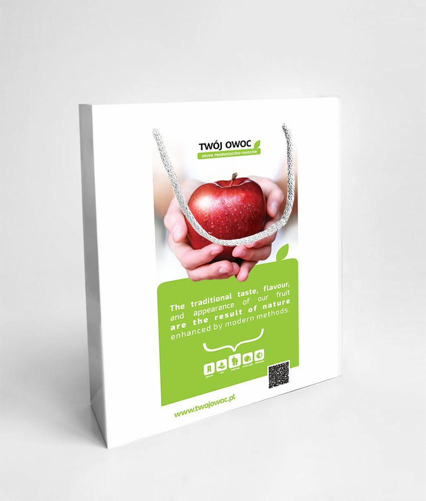 Biała torba papierowa z grafiką jabłka trzymanego w dłoniach, logo 'Twój Owoc' i adresem strony www.twojowoc.pl. Widoczny tekst o smaku i wyglądzie owoców.