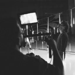 Czarno-białe zdjęcie z planu filmowego: osoba filmująca zza kamery, aktor spoglądający w górę na wiszące lampy w kształcie sopli, rozświetlające przestrzeń.