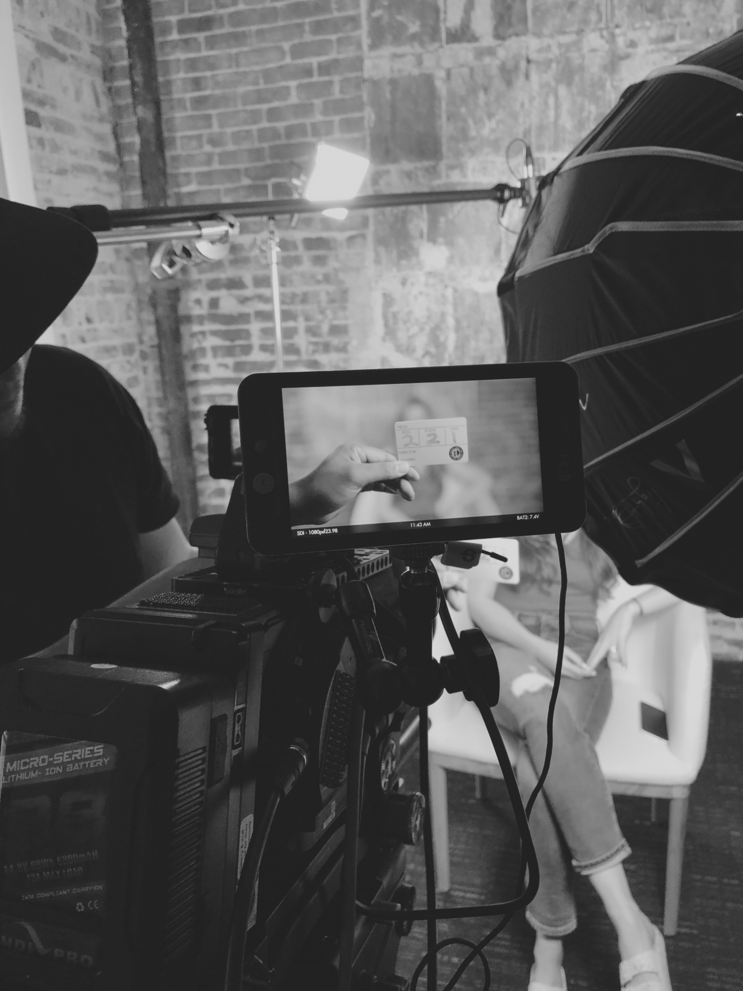 Czarno-białe zdjęcie z planu filmowego: monitor kamery pokazuje zbliżenie na klaps filmowy trzymany w dłoni, w tle oświetlenie studyjne, ceglana ściana i osoba siedząca na białym krześle.