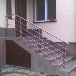 Zewnętrzne schody z szarymi płytkami i metalową balustradą na tle budynku z brązowymi drzwiami i oknem z kwiatami. W tle widoczny pies.