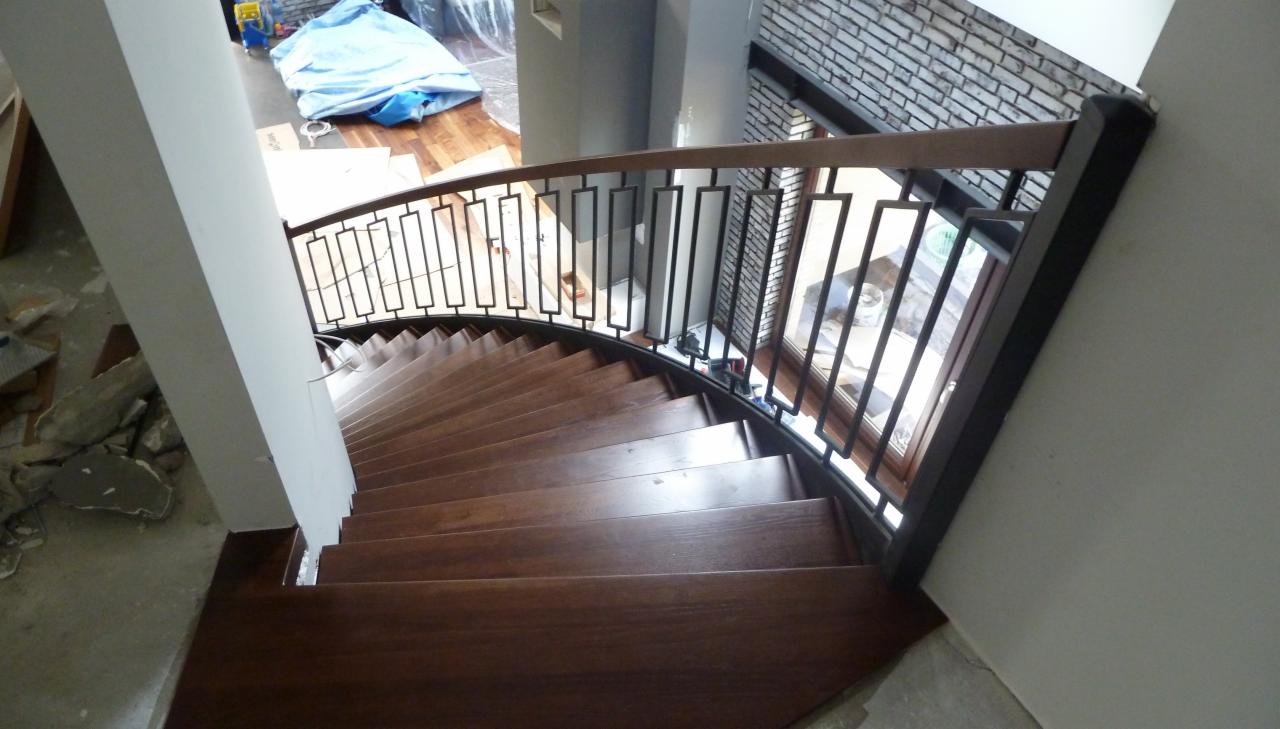 Spiralne schody z ciemnego drewna z metalową balustradą o geometrycznym wzorze, widok z góry, naturalne światło z okna. Częściowo widoczne materiały budowlane.