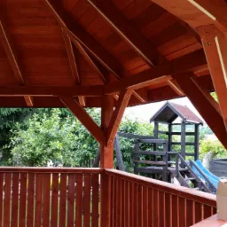 Drewniana altana z widoczną konstrukcją dachu i balustradą, w tle plac zabaw i zieleń. Elementy drewniane w ciepłym, brązowym odcieniu. Widok z wnętrza altany.