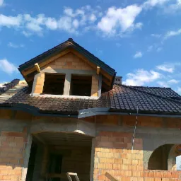 Wznoszenie domu z cegły: częściowo pokryty dach z ciemnej dachówki ceramicznej, widoczne okna poddasza i fragment błękitnego nieba z chmurami.