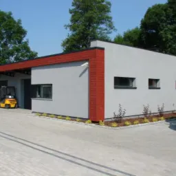 Szary budynek magazynowy z czerwonymi elementami wykończenia, widoczny wózek widłowy i samochód dostawczy na kostce brukowej, otoczony zielenią.