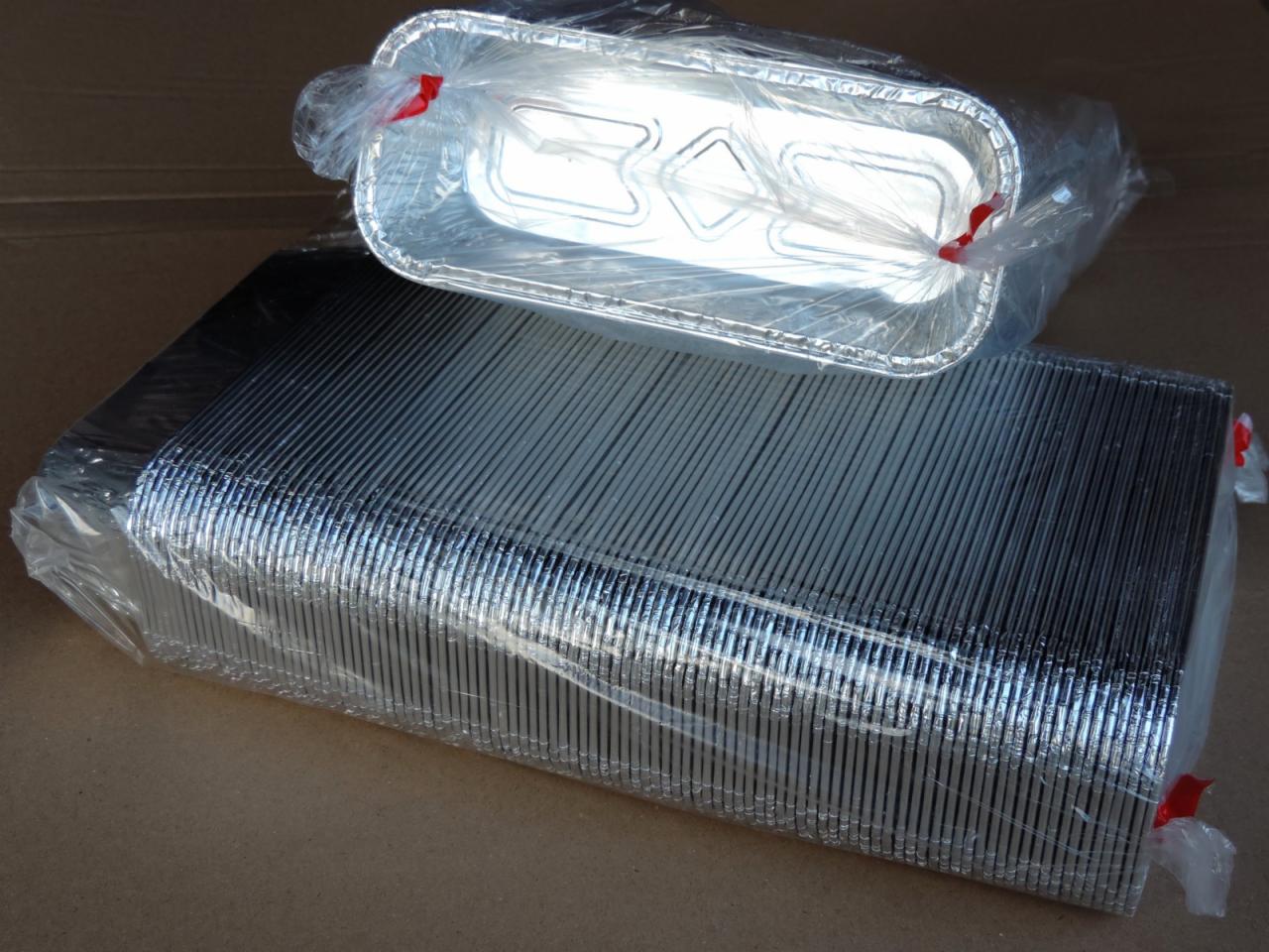 Prostokątne aluminiowe foremki do pieczenia, zapakowane w folię, ułożone w dwie paczki. Jedna paczka zawiera pojedynczą foremkę, druga duży stos foremek.