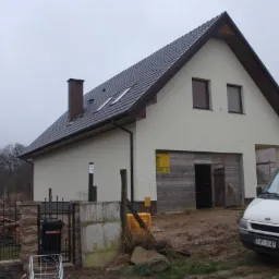 Nowo otynkowany dom jednorodzinny z ciemną dachówką, widoczne fragmenty drewnianej konstrukcji garażu i biały bus firmy budowlanej na błotnistym podwórku.