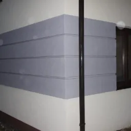 Fragment ocieplonej elewacji budynku z szarymi panelami izolacyjnymi, widoczny narożnik i okno z brązową ramą, rura spustowa na pierwszym planie.