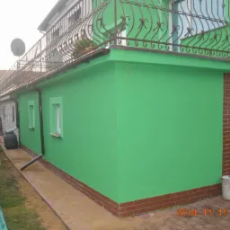 Świeżo pomalowana, intensywnie zielona elewacja domu z brązowymi detalami (drzwi, cokół z cegły) i metalową balustradą na balkonie. Widoczny kubeł po farbie.