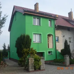 Dom z zieloną elewacją, widok z zewnątrz, kostka brukowa na podjeździe, drzewa iglaste. Dom po remoncie z nową, pomalowaną elewacją na jaskrawy kolor.