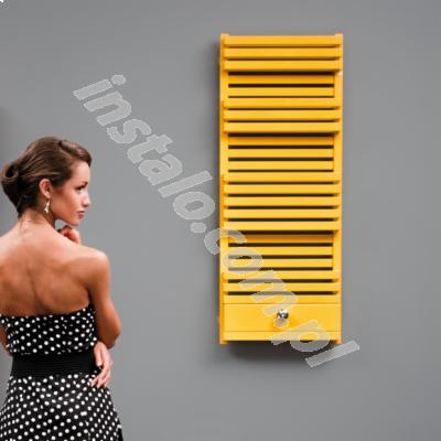 Żółty, dekoracyjny grzejnik łazienkowy na szarym tle, z kobietą w sukience w groszki, patrzącą w lewo. Grzejnik z poziomymi szczebelkami i małą szafką.