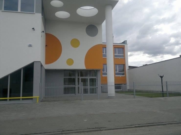 Nowoczesny budynek z szarą metalową bramą i kolorowymi okrągłymi dekoracjami na elewacji. Widoczne okna i oświetlenie.