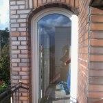 Łukowe okno PCV w murze z cegły klinkierowej, widok z zewnątrz. Nietypowy kształt okna w budynku z ceglaną elewacją.