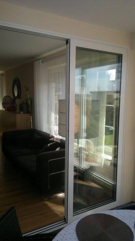 Nowoczesne, przesuwne drzwi balkonowe z białymi ramami, otwierające się na jasny salon. Widoczny fragment mebli i dekoracje wnętrza.